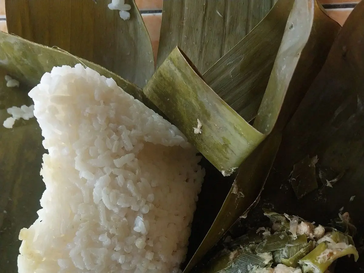 Arroz blanco junto a un paquete abierto de hoja de plátano con botok de coco y hierbas