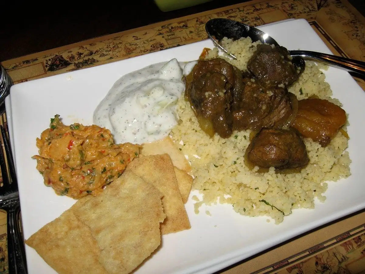 Plato blanco con cuscús, carne especiada guisada, yogur, puré de verduras y crujientes de pan