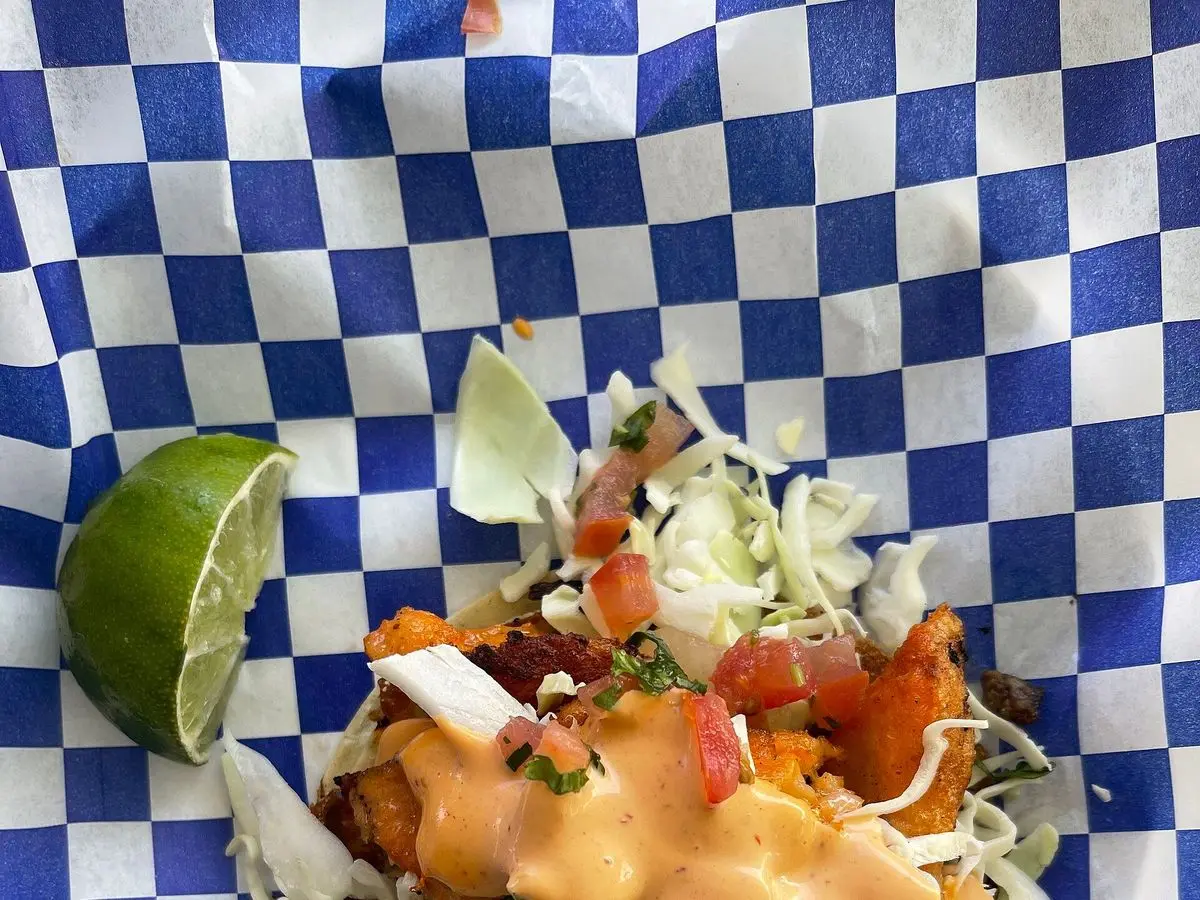 Taco con repollo en tiras, salsa cremosa anaranjada y lima sobre un mantel de cuadros azules