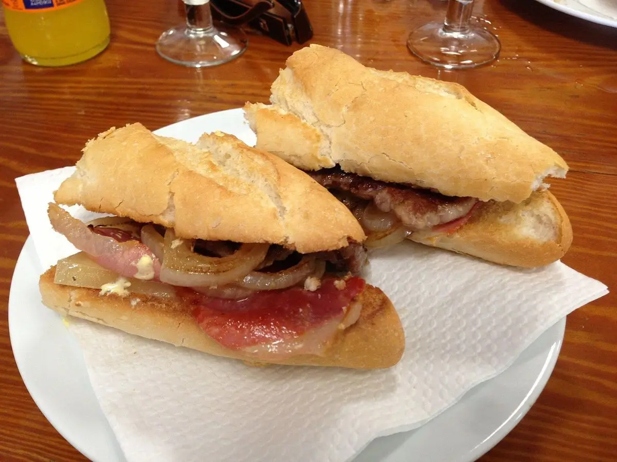 Bocadillo de pan crujiente partido en dos relleno de carne, jamón y cebolla sobre una servilleta