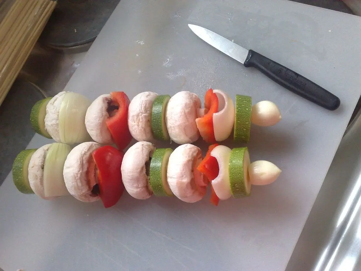 Brochetas de verduras con calabacín, champiñón, pimiento rojo y cebolla ensartadas sobre una tabla junto a un cuchillo