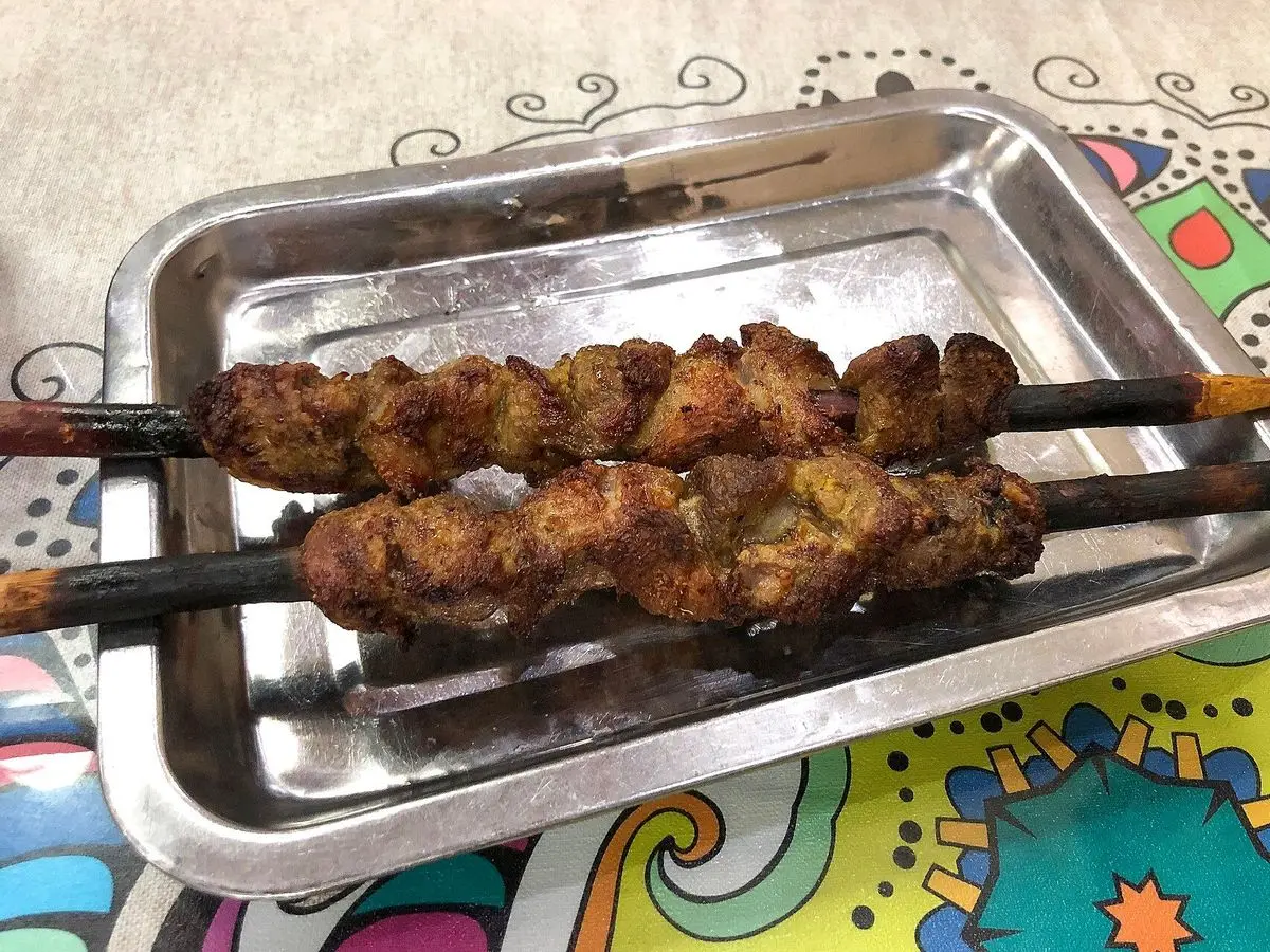 Dos brochetas finas de cordero especiado a la parrilla en una bandeja metálica sobre un mantel de colores