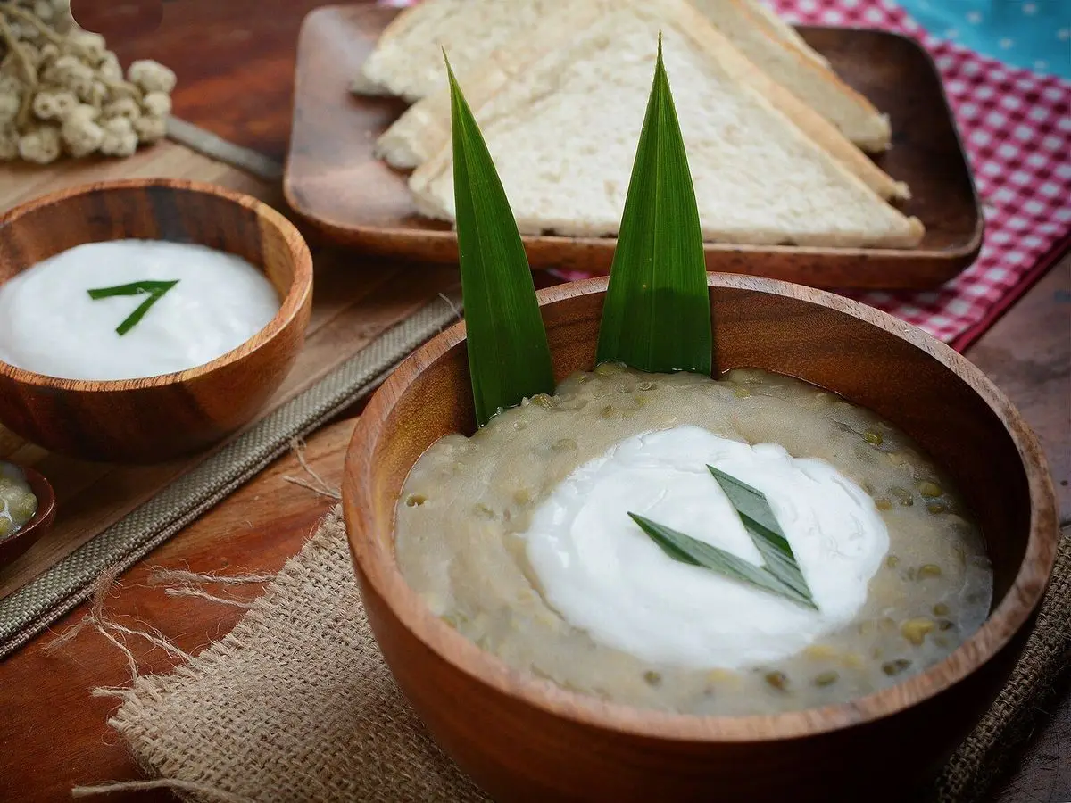 Cuencos de madera con gachas de judía mungo cubiertas de leche de coco y hojas de pandan, con pan al fondo