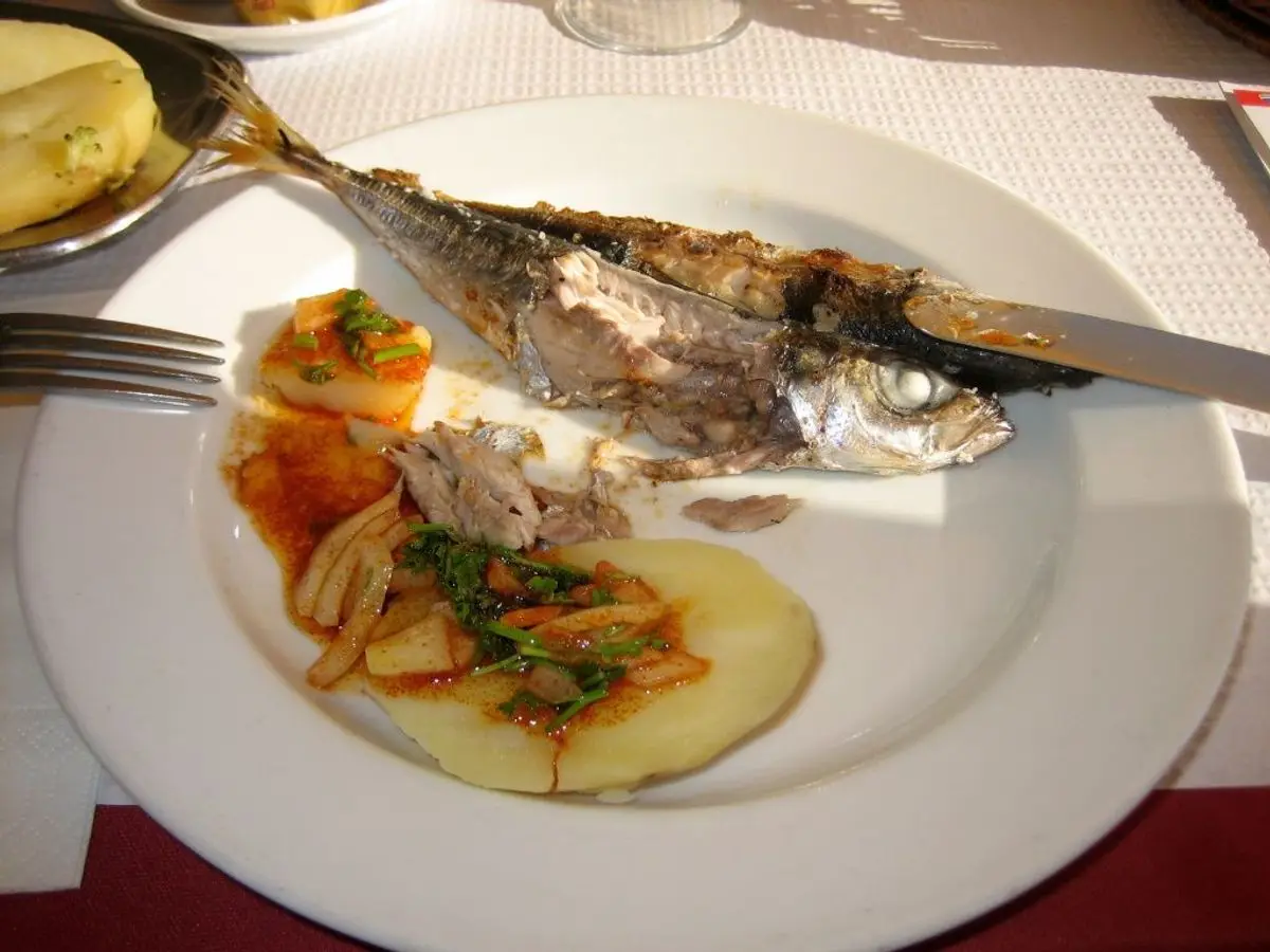 Pescado azul entero a la plancha sobre un lecho de patata, cebolla y tomate con hierbas en un plato blanco