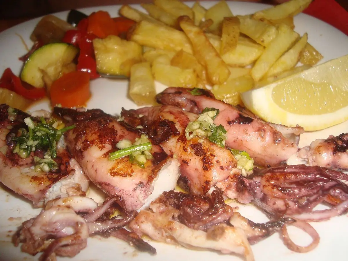 Calamares a la plancha con ajo y perejil, patatas fritas, limón y verduras asadas en un plato blanco