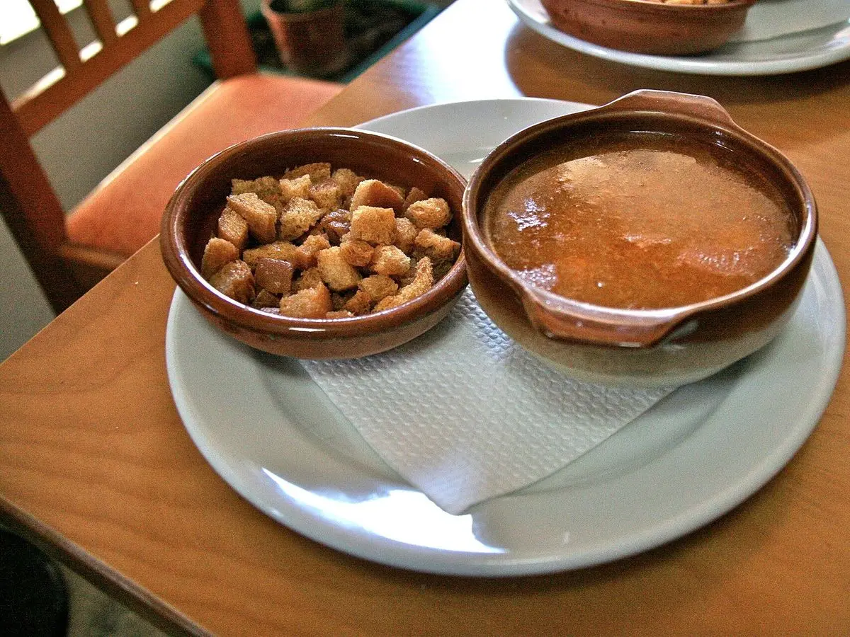 Cazuela de barro con sopa de pescado y marisco junto a un cuenco de picatostes de pan