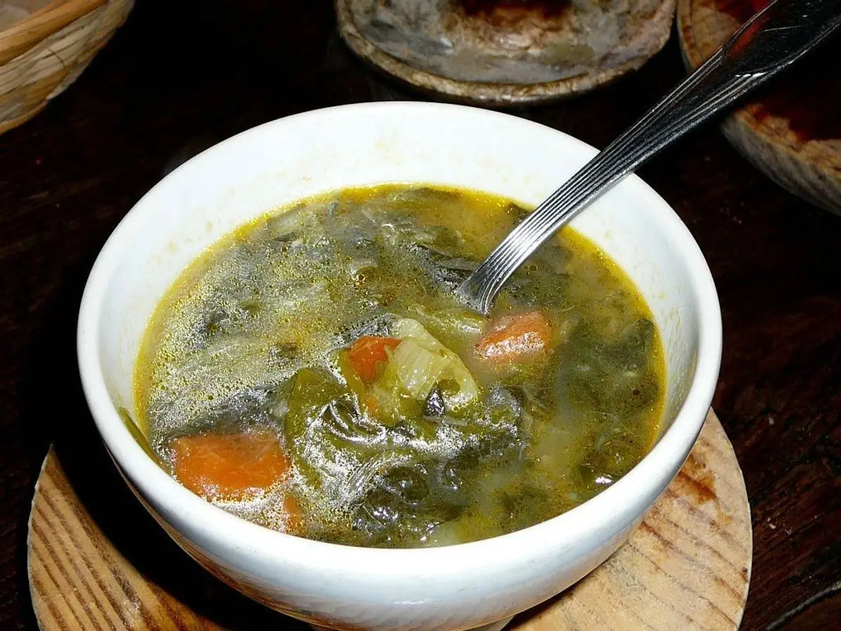 Cuenco blanco de caldo con hojas de berza, patata, zanahoria y trozos de carne, con cuchara