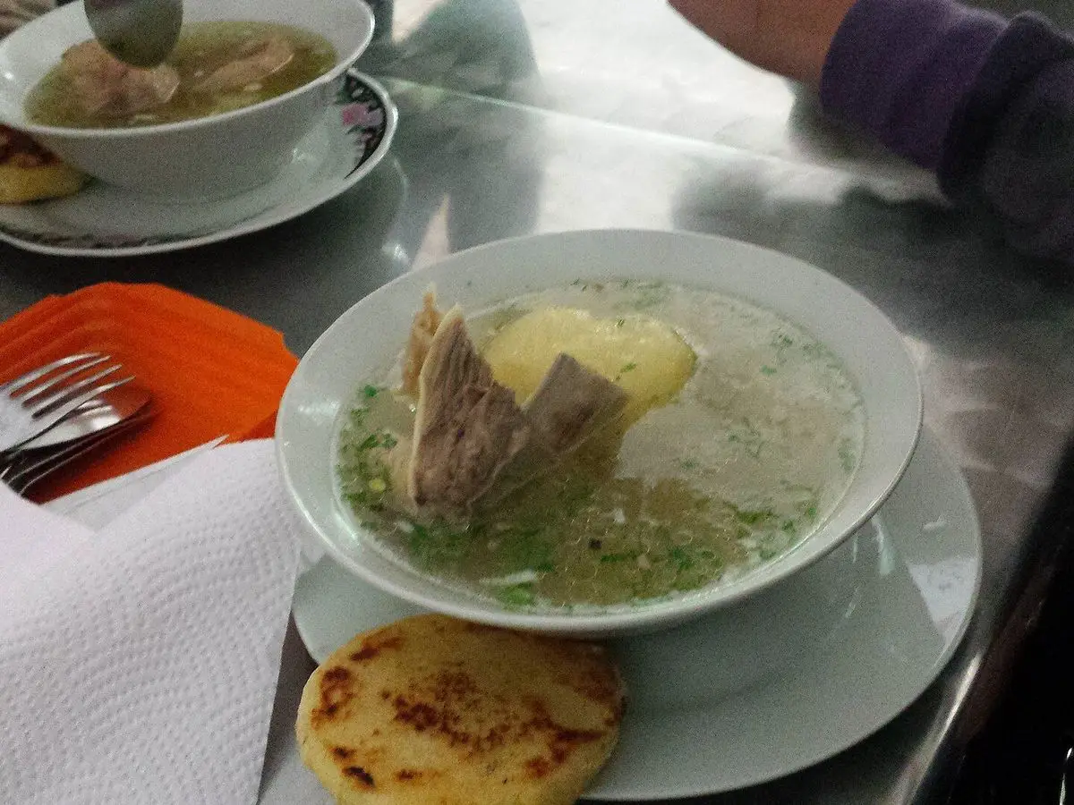 Plato de caldo de costilla con patata y hierbas, acompañado de una arepa