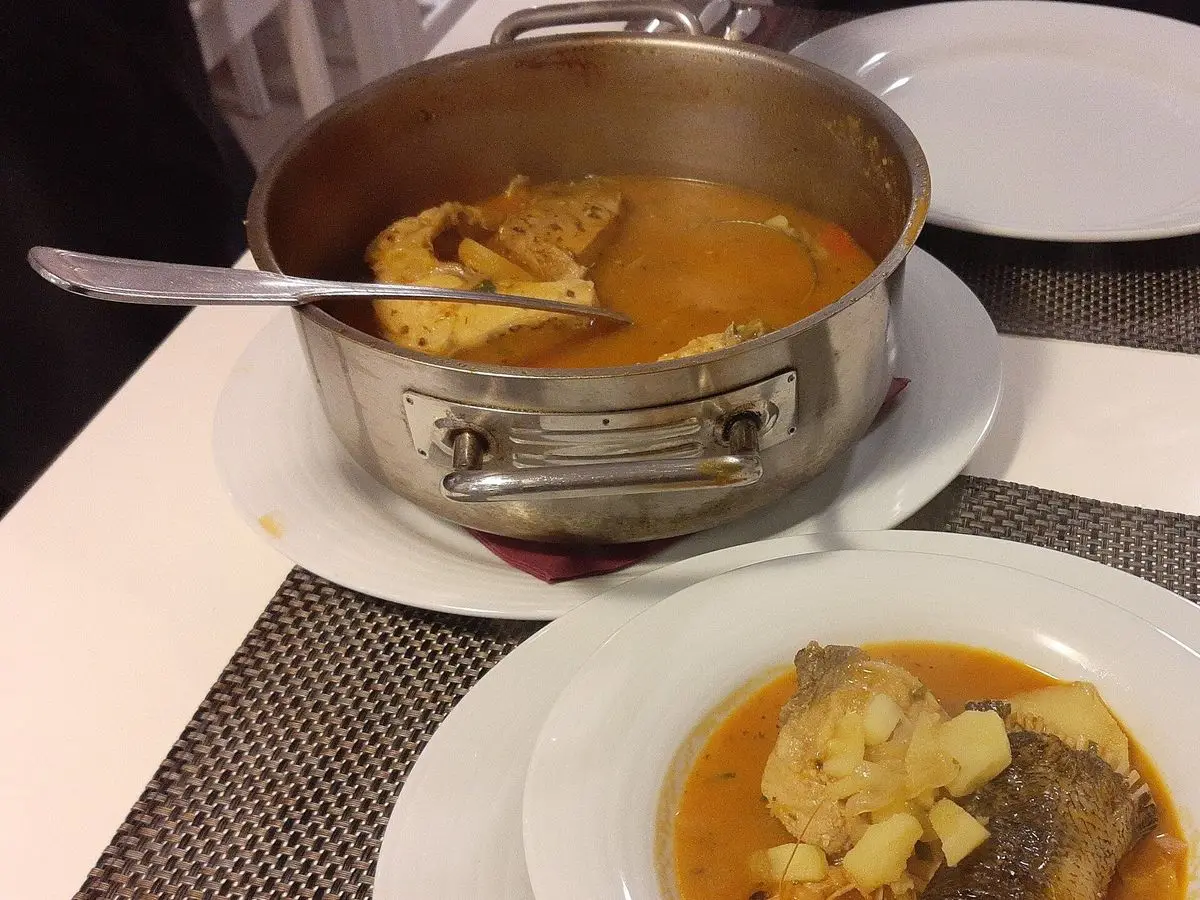 Sopa de pescado anaranjada con patata y trozo de pescado en un plato, con olla al fondo