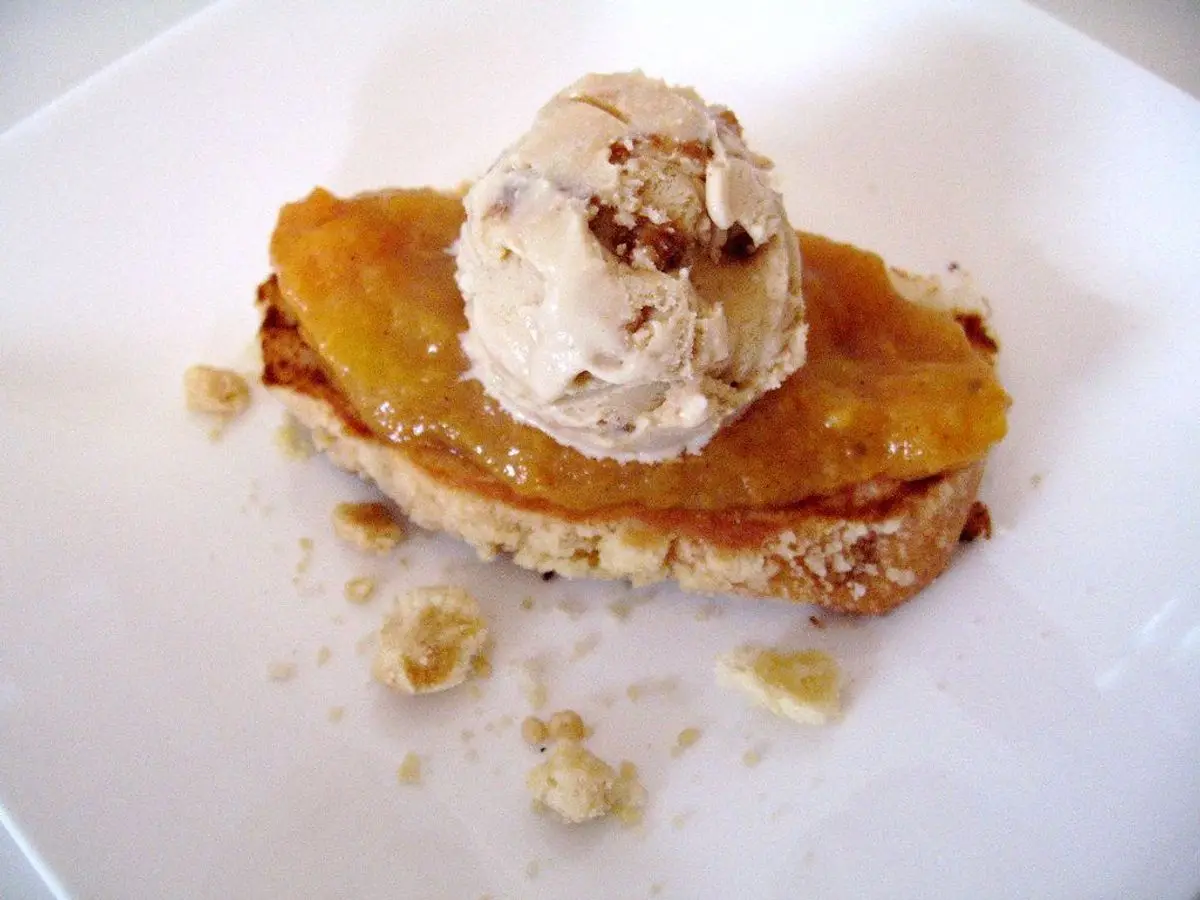 Fruta anaranjada asada y caramelizada sobre una base dorada con una bola de crema helada encima en un plato blanco