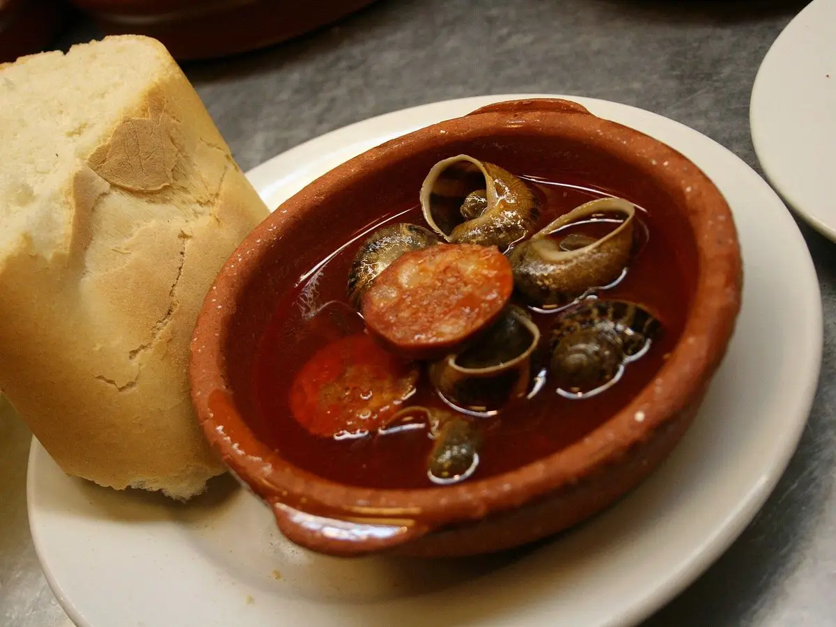 Cazuela de barro con caracoles en salsa roja y rodajas de chorizo junto a un trozo de pan