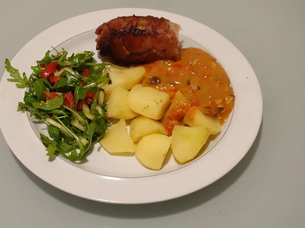 Plato blanco con albóndiga de carne, patatas cocidas en salsa y ensalada de hojas verdes y tomate