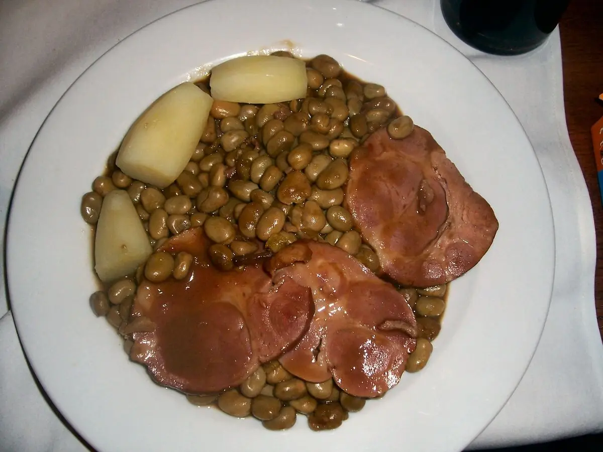 Plato blanco con lonchas de cerdo ahumado sobre habas guisadas y patatas cocidas