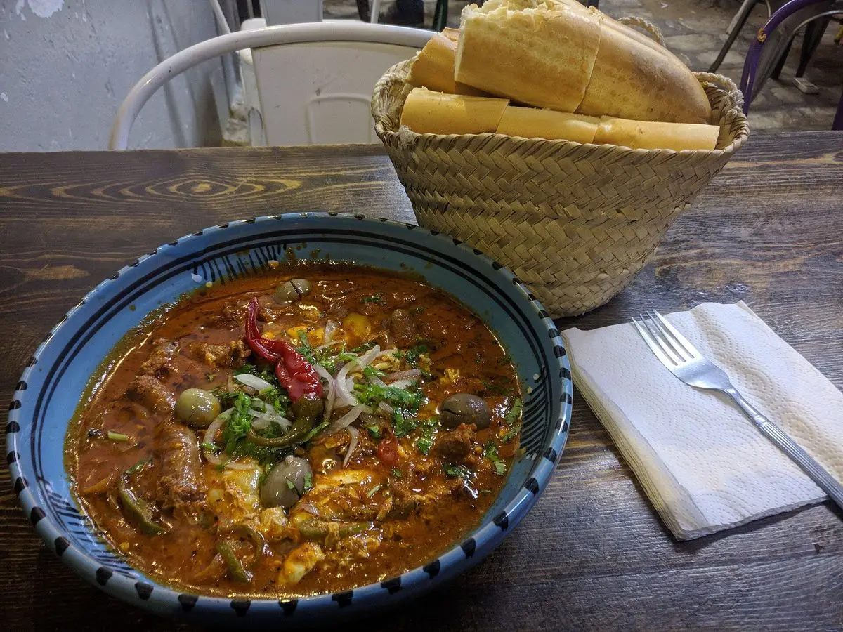 Cuenco azul de chakchouka con salsa de tomate, huevo, guindilla y hierbas junto a una cesta de pan