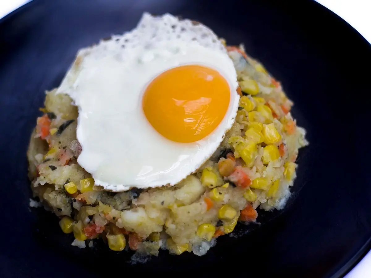 Plato oscuro con puré de verduras con maíz y guisantes coronado por un huevo frito