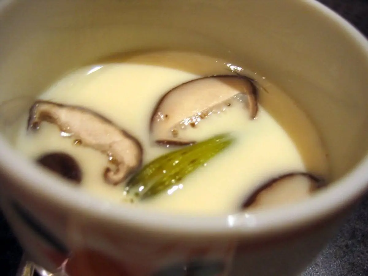 Flan japonés de huevo cremoso con setas shiitake y tallo verde servido en un cuenco de cerámica blanco
