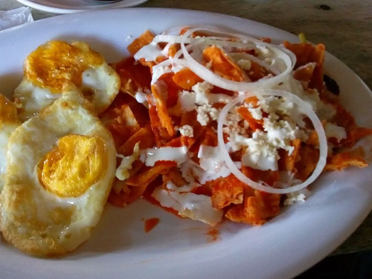 Chilaquiles en salsa roja con huevos fritos, cebolla en aros y queso fresco desmenuzado en un plato blanco
