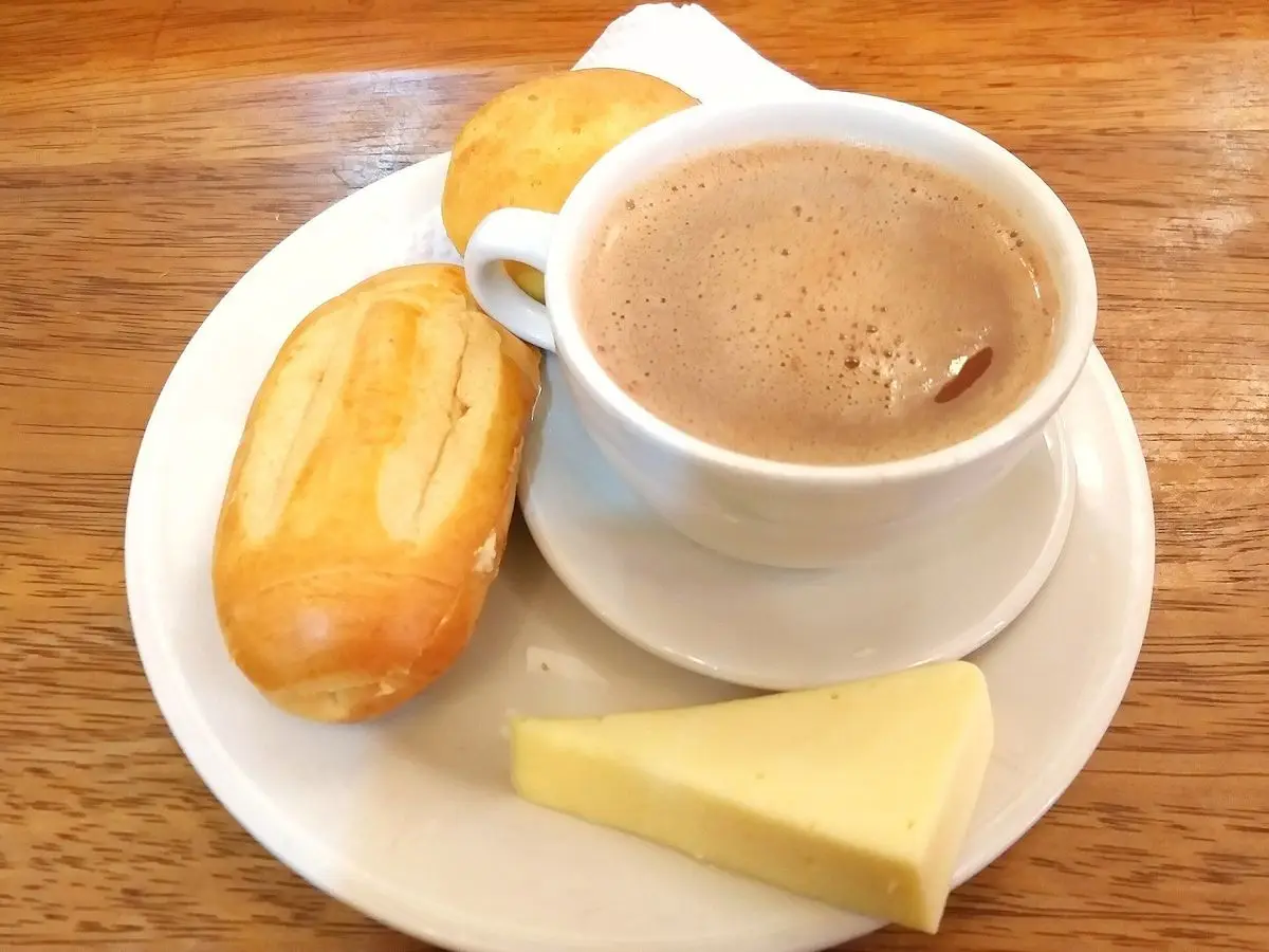 Taza de chocolate caliente con un panecillo y una cuña de queso fresco sobre un plato blanco