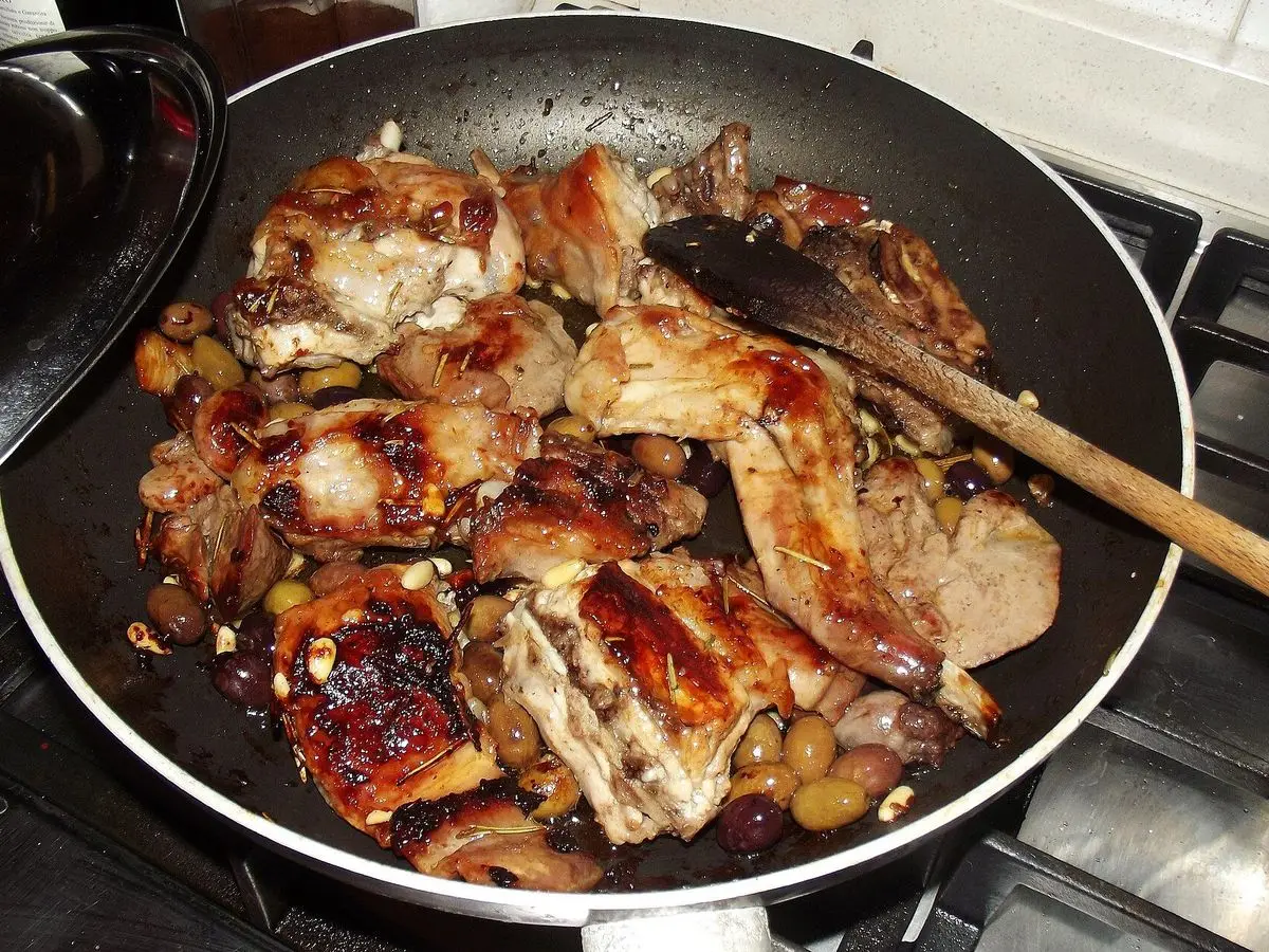 Sartén con trozos de conejo dorados guisados con aceitunas y piñones y una cuchara de madera