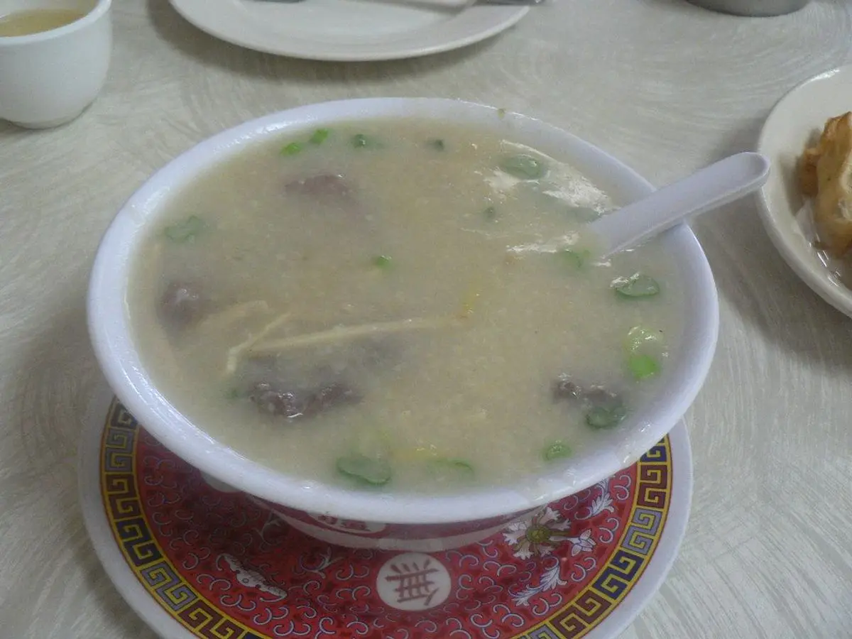 Cuenco blanco de congee, sopa espesa de arroz con verduras verdes y trozos de proteína, con cuchara