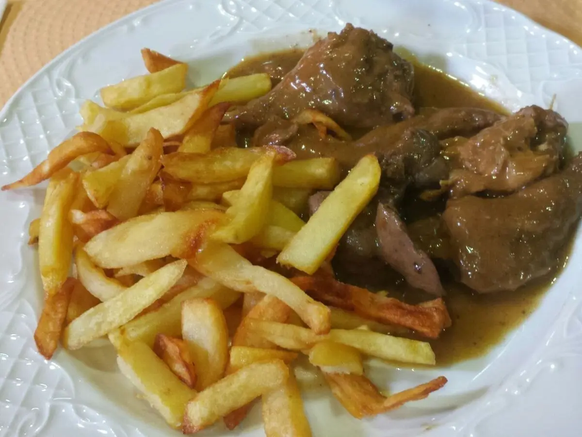 Plato de trozos de conejo estofado en salsa oscura acompañados de patatas fritas