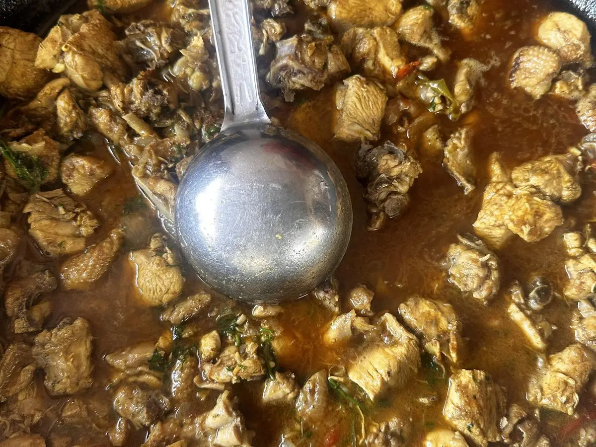 Trozos de pollo guisados en su caldo dentro de una sartén de hierro, con un cucharón