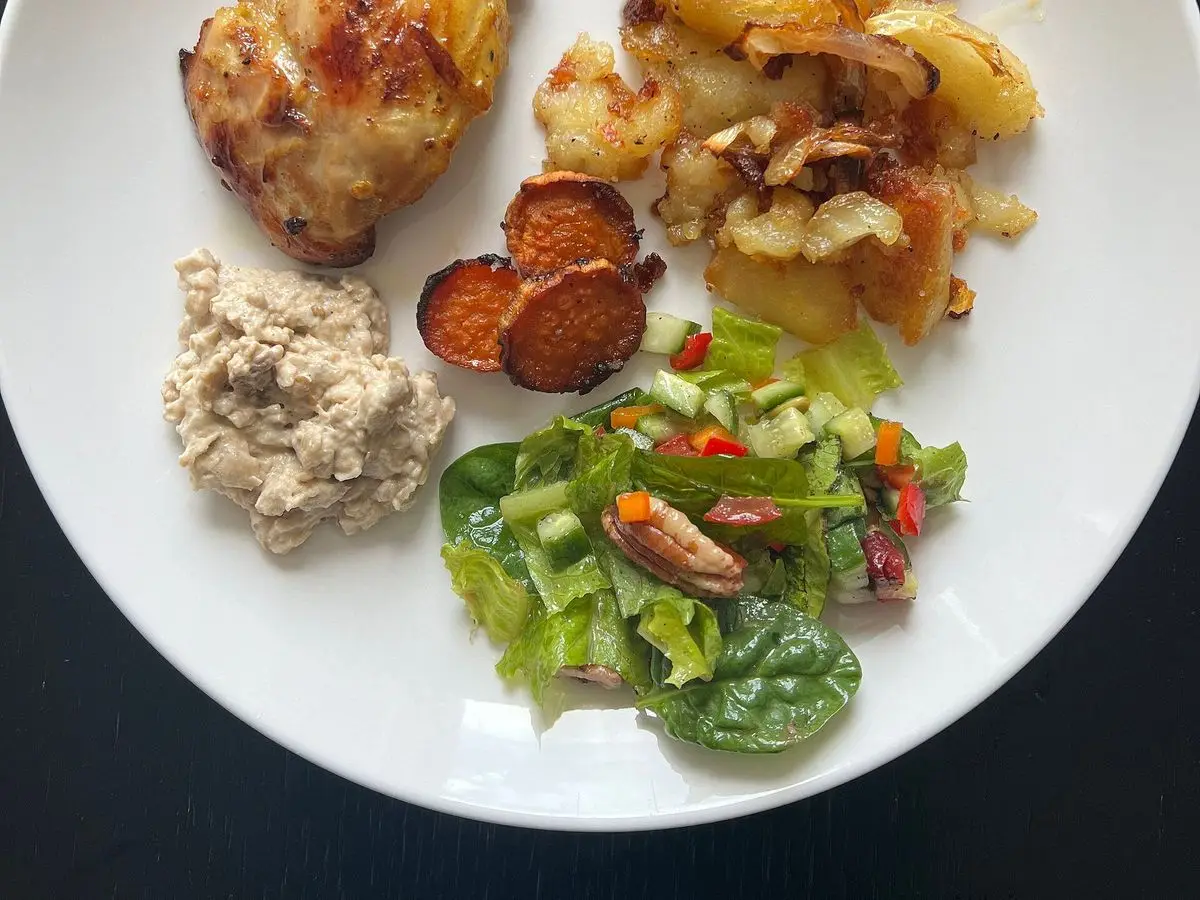 Plato blanco con pollo asado dorado, patatas asadas, rodajas de boniato y ensalada verde