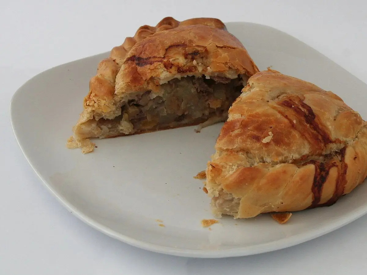 Empanada tipo Cornish pasty de masa dorada cortada por la mitad sobre un plato blanco, con relleno de carne y patata