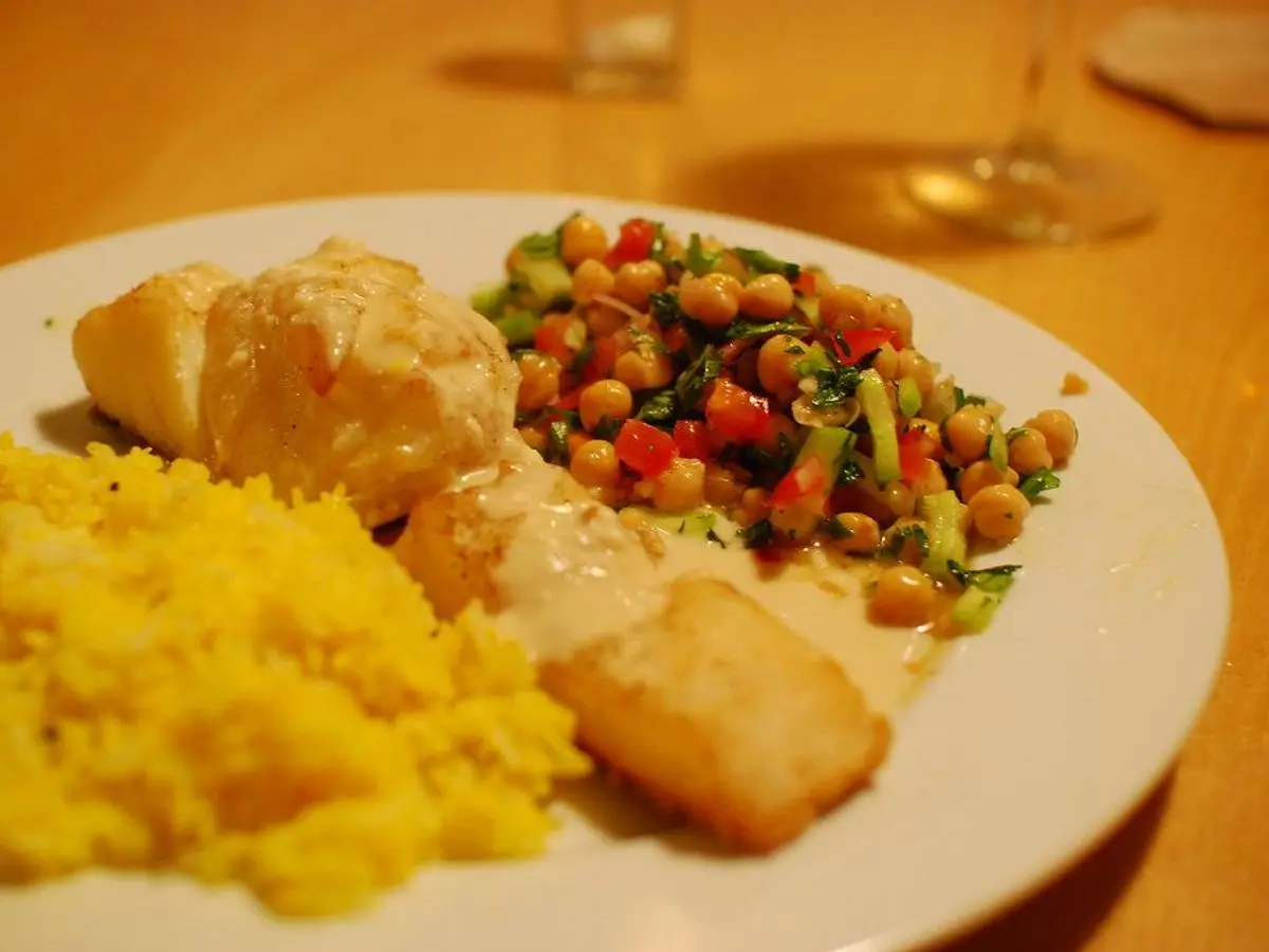 Bacalao horneado con salsa de tahini, ensalada de garbanzos con tomate y arroz amarillo en un plato blanco
