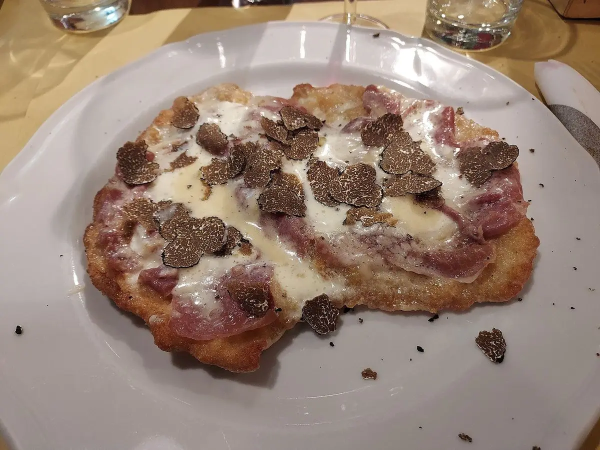 Cotoletta empanada a la boloñesa cubierta de queso fundido, jamón y láminas de trufa en un plato blanco
