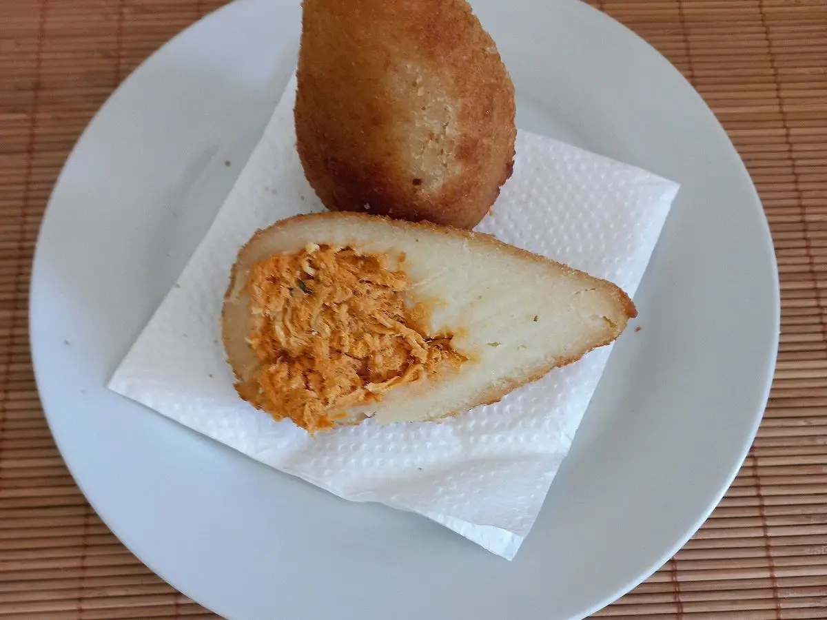 Coxinha brasileña empanada, una de ellas abierta mostrando el relleno de pollo desmenuzado, en un plato blanco