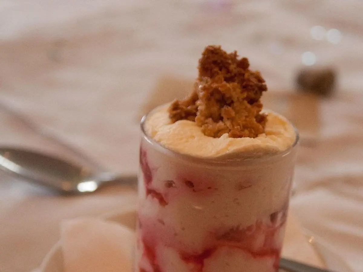 Vaso con capas de crema y frambuesa coronado con avena tostada, junto a una cucharilla