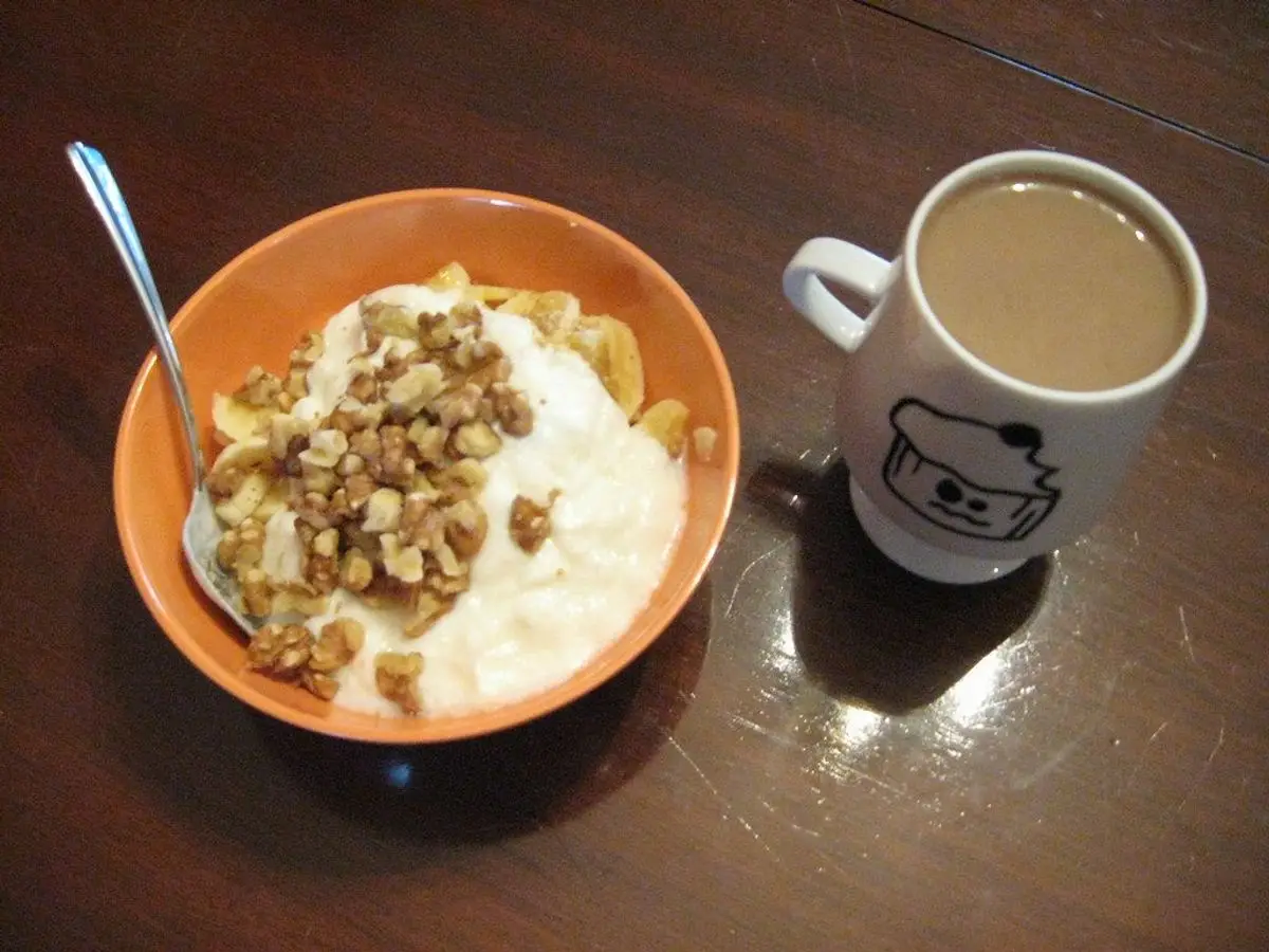 Cuenco naranja de yogur cremoso con plátano en rodajas y nueces troceadas junto a una taza de café