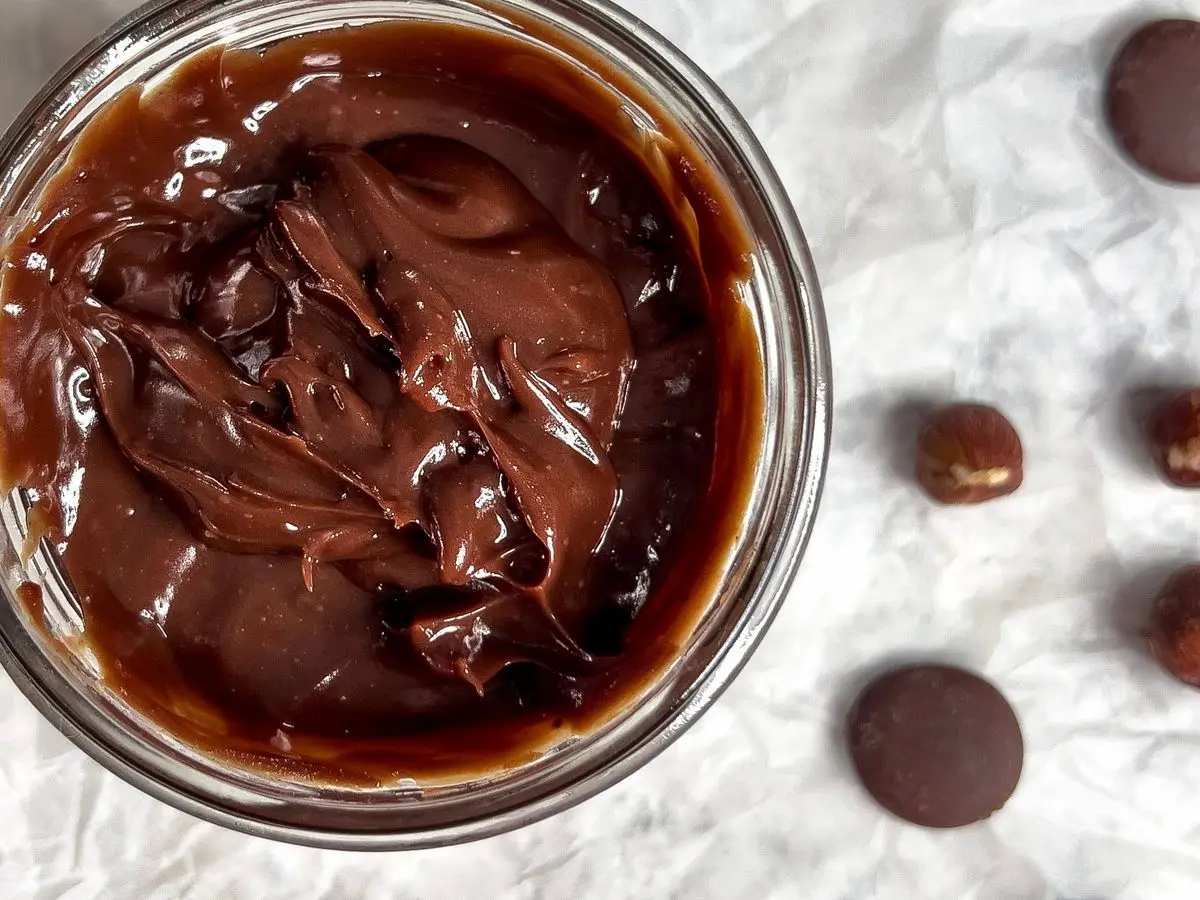 Tarro de cristal con crema de chocolate y avellanas, junto a avellanas y pepitas de chocolate