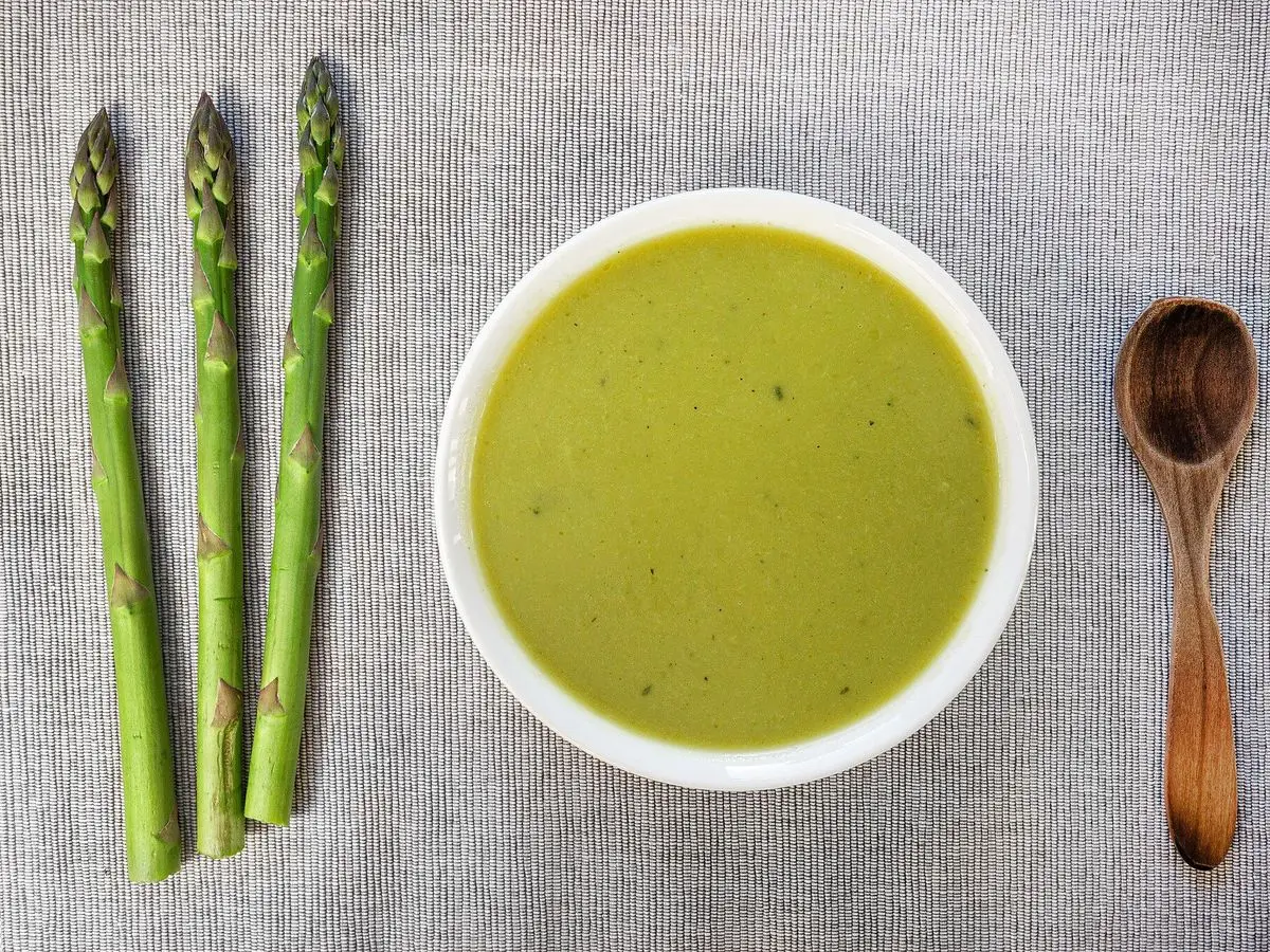 Cuenco blanco de crema verde de espárragos junto a espárragos verdes frescos y una cuchara de madera