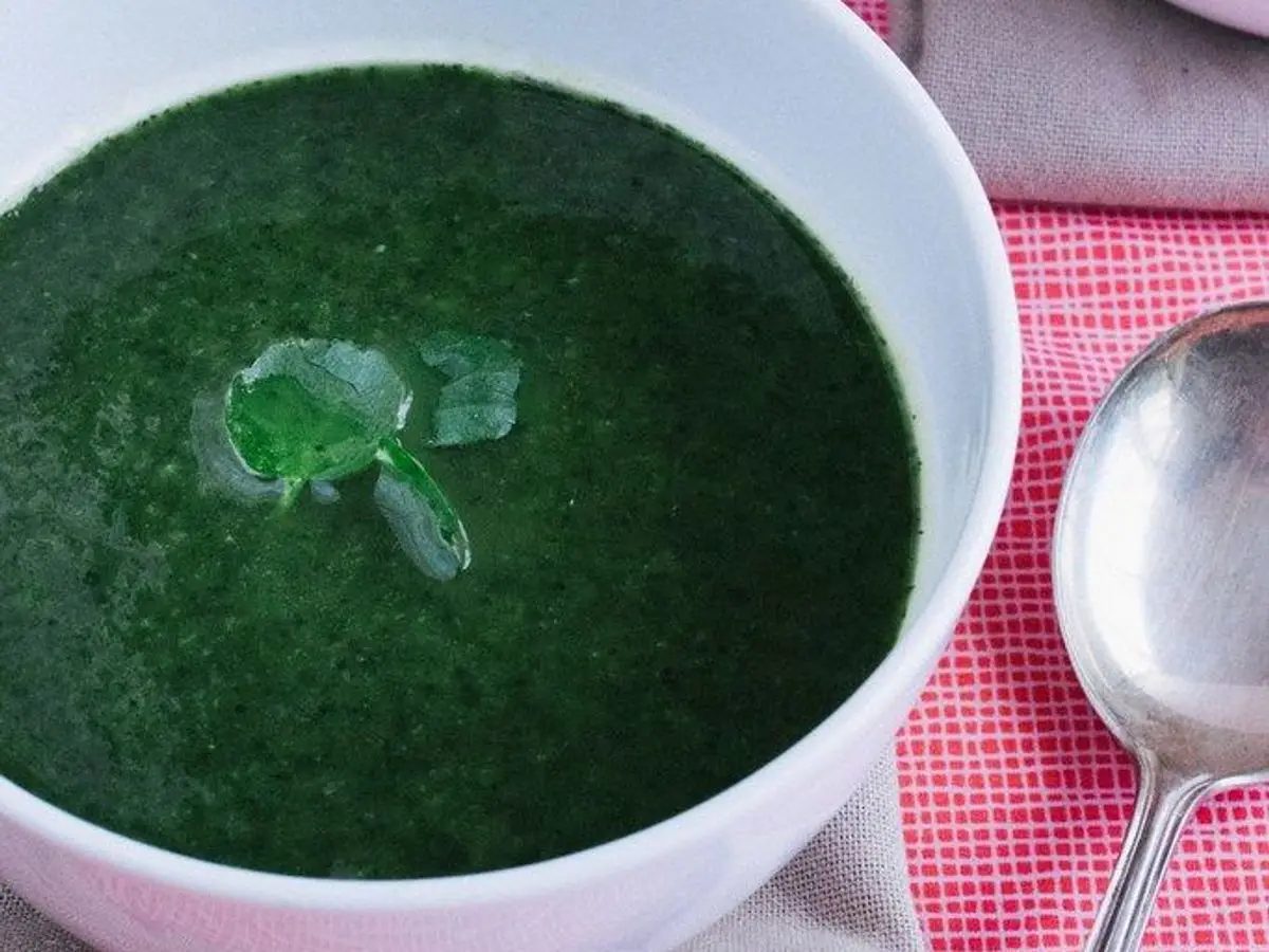 Cuenco blanco de crema verde de espinacas con una hoja por encima, junto a una cuchara