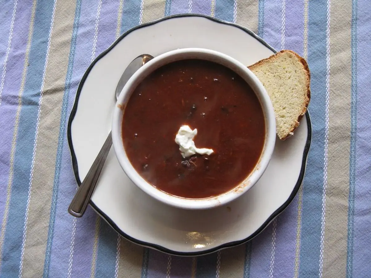 Taza de crema de frijoles negros con una cucharada de crema agria, cuchara y pan sobre un mantel de rayas