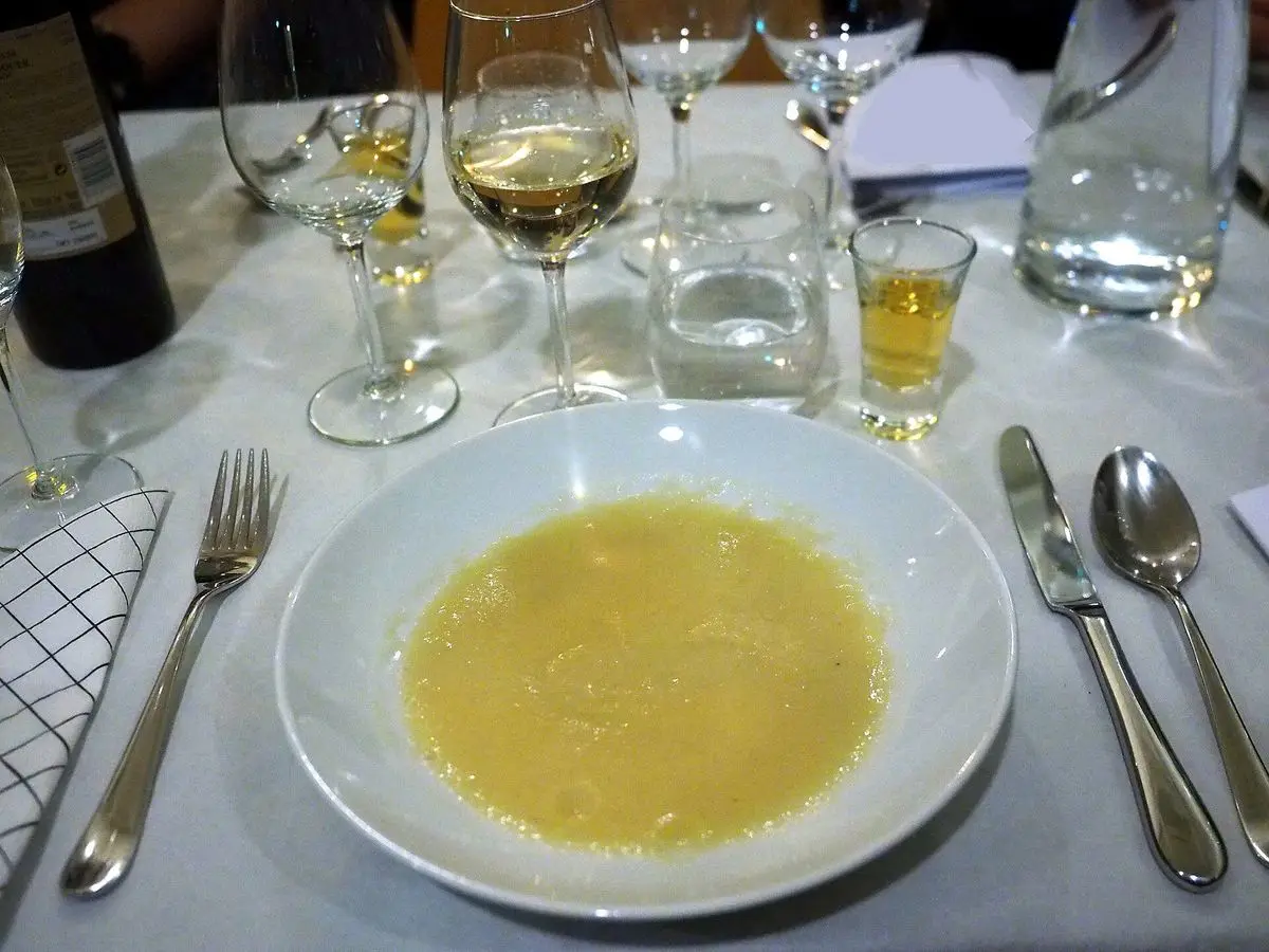 Plato hondo blanco de crema amarilla de tupinambo sobre una mesa puesta con copa de vino