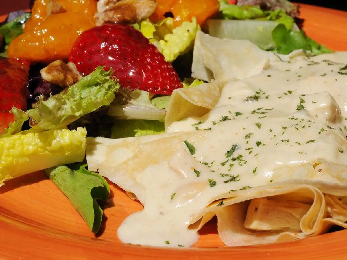 Crepes rellenos cubiertos de salsa cremosa con hierbas, junto a una ensalada de fresas y naranja
