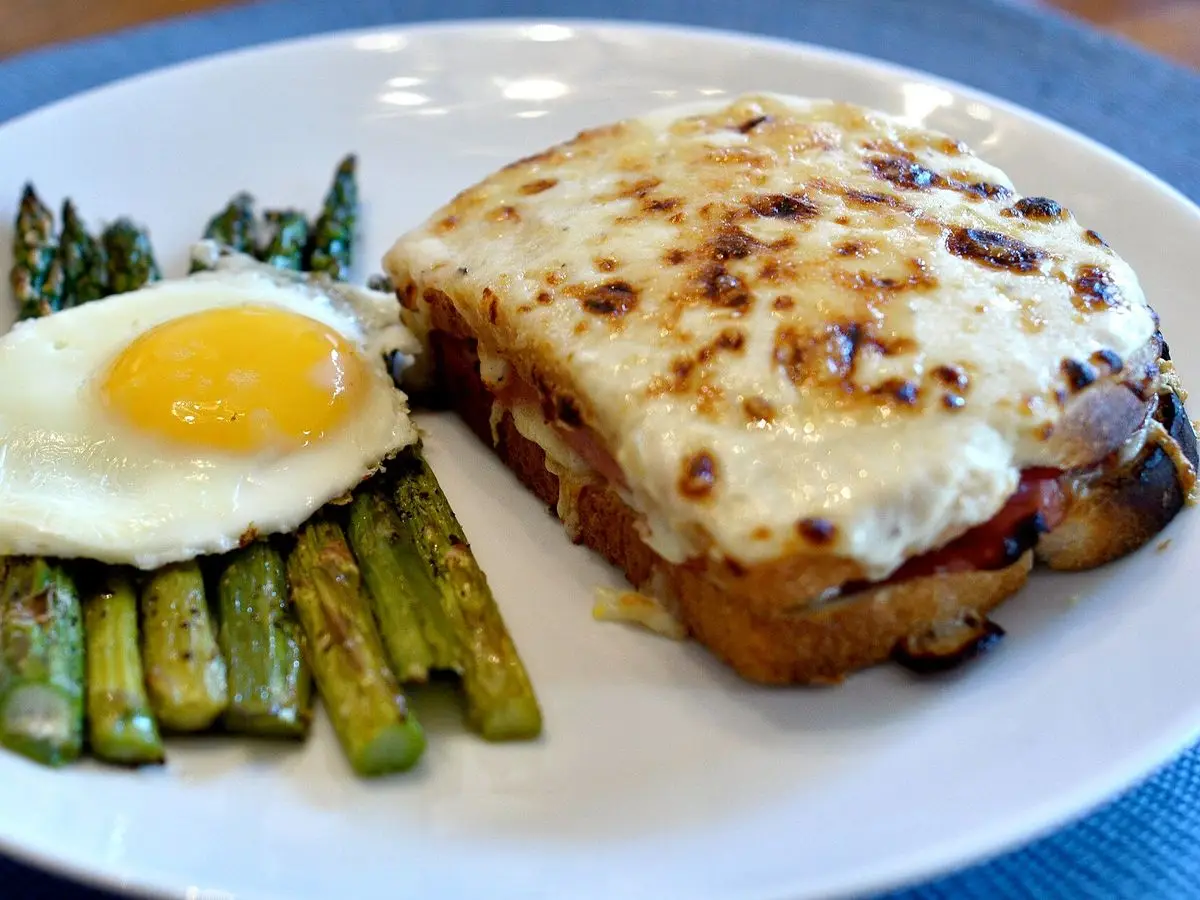 Sándwich gratinado de jamón y queso con huevo frito y espárragos verdes en un plato blanco