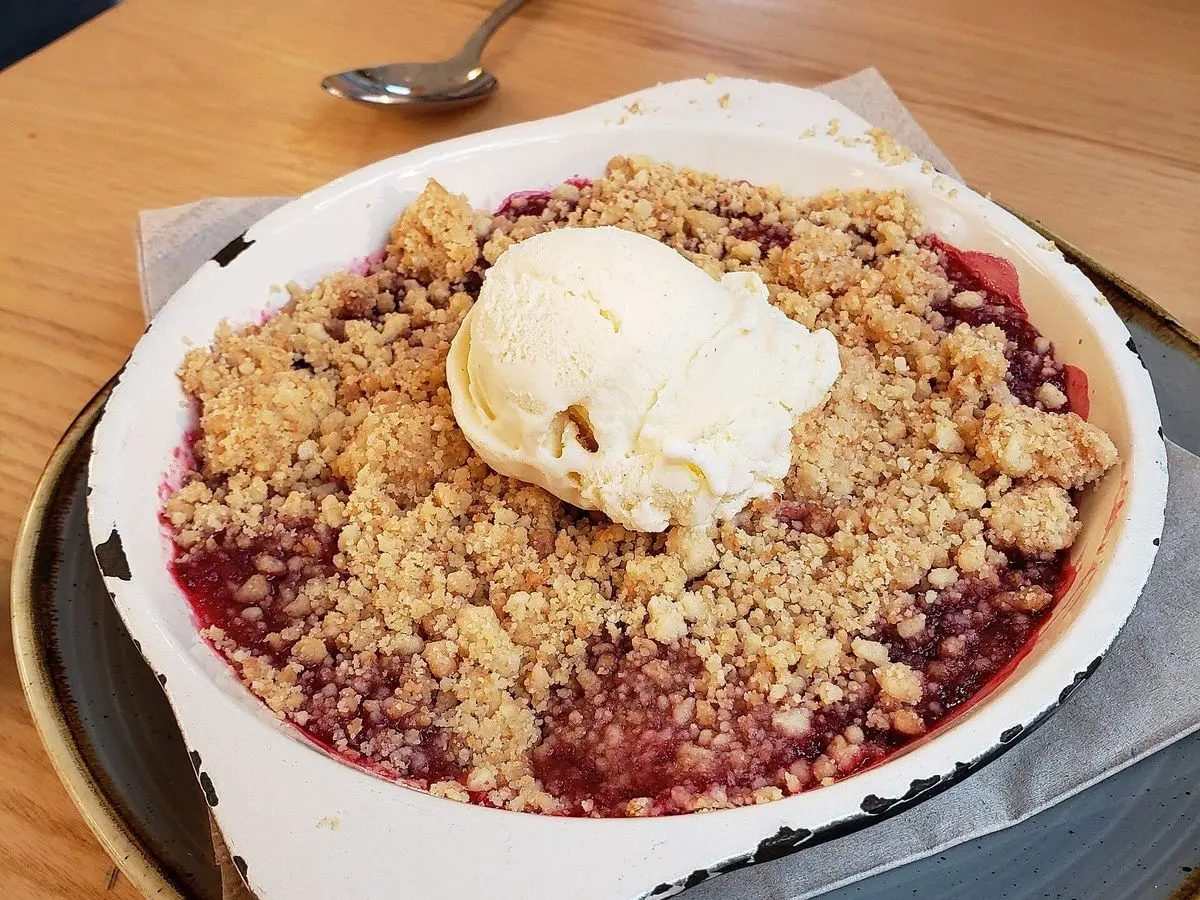 Crumble de frutos rojos con cobertura crujiente de avena y una bola cremosa blanca encima, en un plato