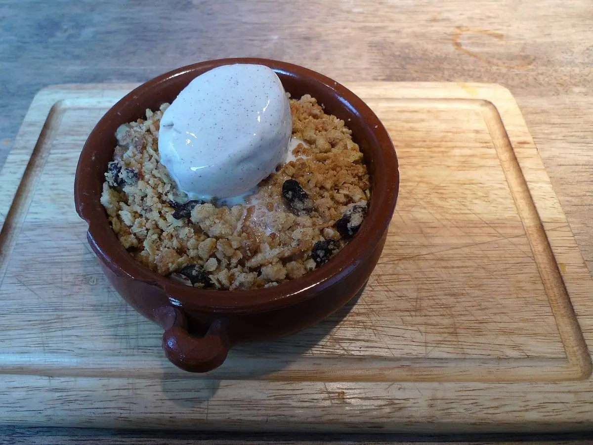 Cazuelita de barro con crumble de manzana y pera y una bola de helado, sobre una tabla de madera