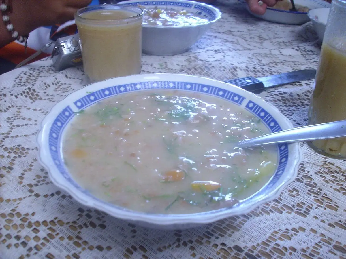 Plato hondo de sopa espesa de trigo con verduras sobre un mantel de encaje y un vaso de jugo