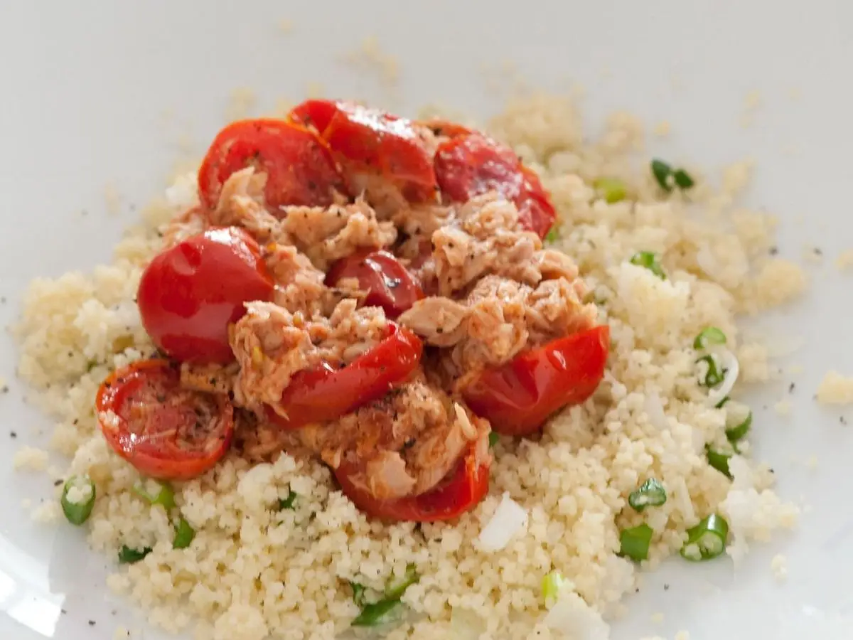 Cuscús con pescado en conserva desmigado, tomates cherry asados y cebolleta picada en un plato blanco