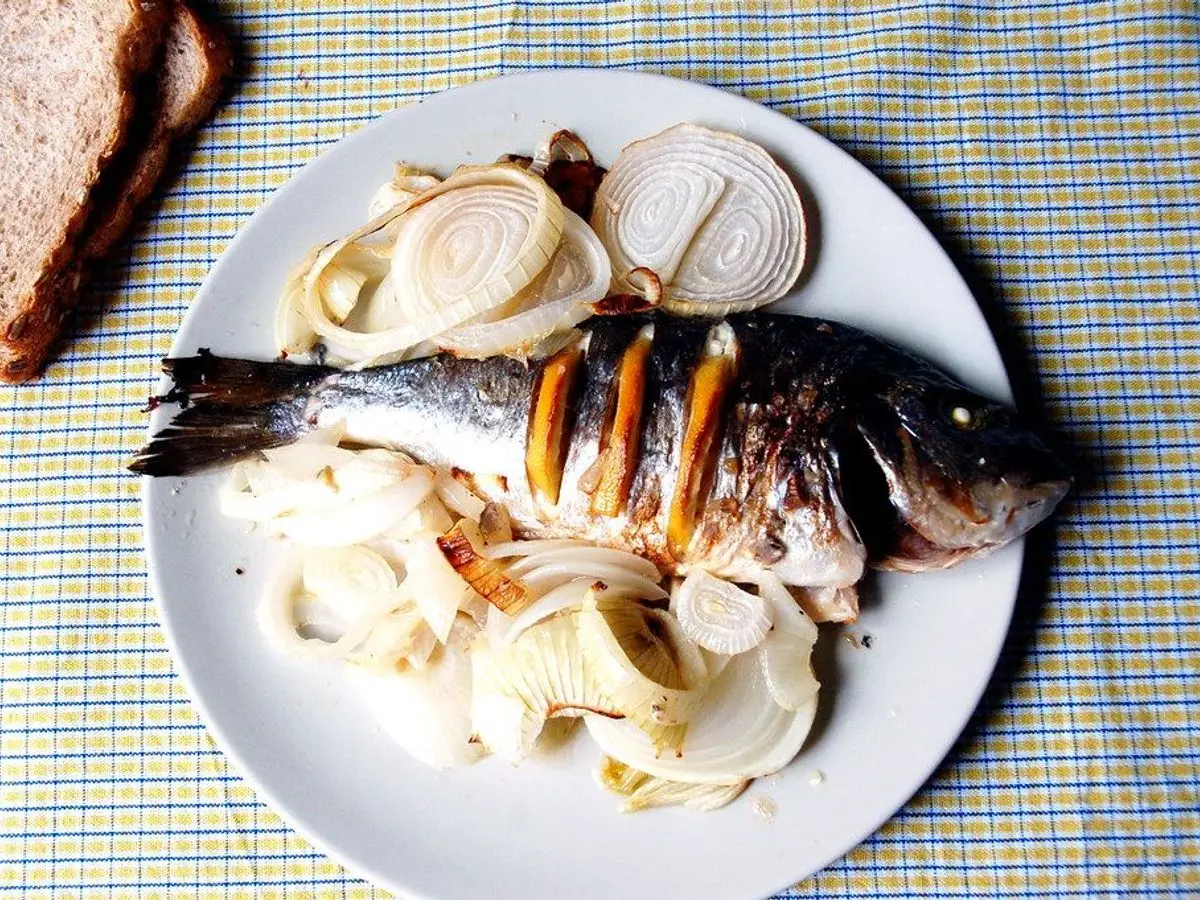 Pescado entero al horno sobre cebolla y rodajas de limón en un plato blanco, con pan al lado