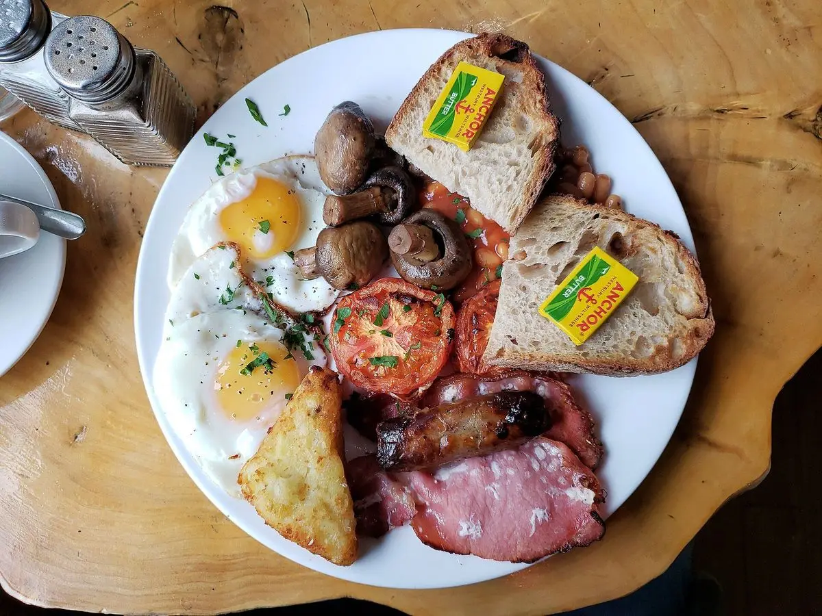 Plato de desayuno inglés con huevos fritos, bacon, champiñones, tomate y rebanadas de pan sobre una mesa de madera