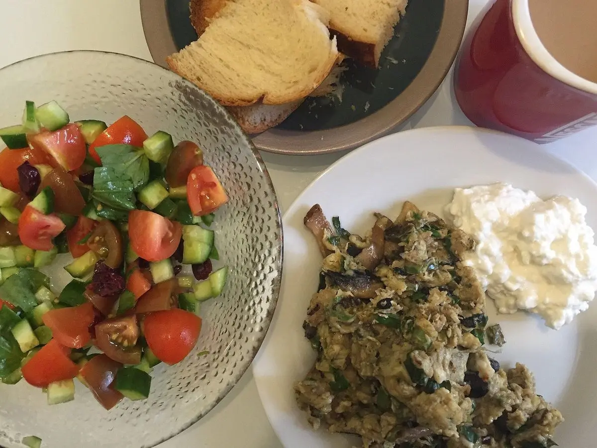 Desayuno con ensalada picada de tomate y pepino, queso fresco blanco, pan y huevos revueltos con setas y hierbas