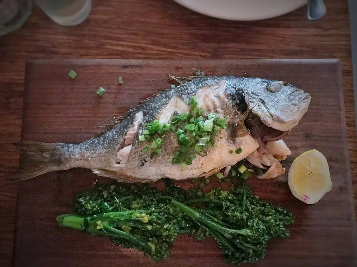 Dorada entera asada sobre una tabla de madera con hierbas picadas y verduras verdes