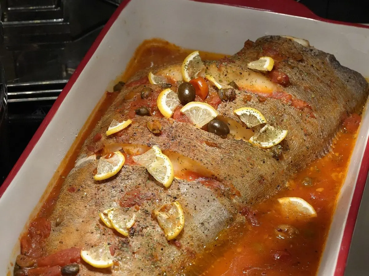 Pescado entero al horno en una fuente blanca con salsa de tomate y rodajas de limón