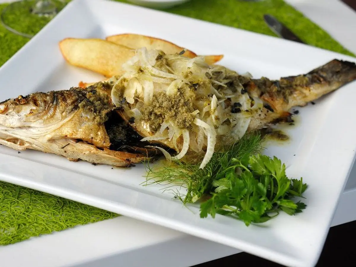 Pescado entero asado con salsa verde, cebolla en juliana y hierbas frescas sobre plato blanco